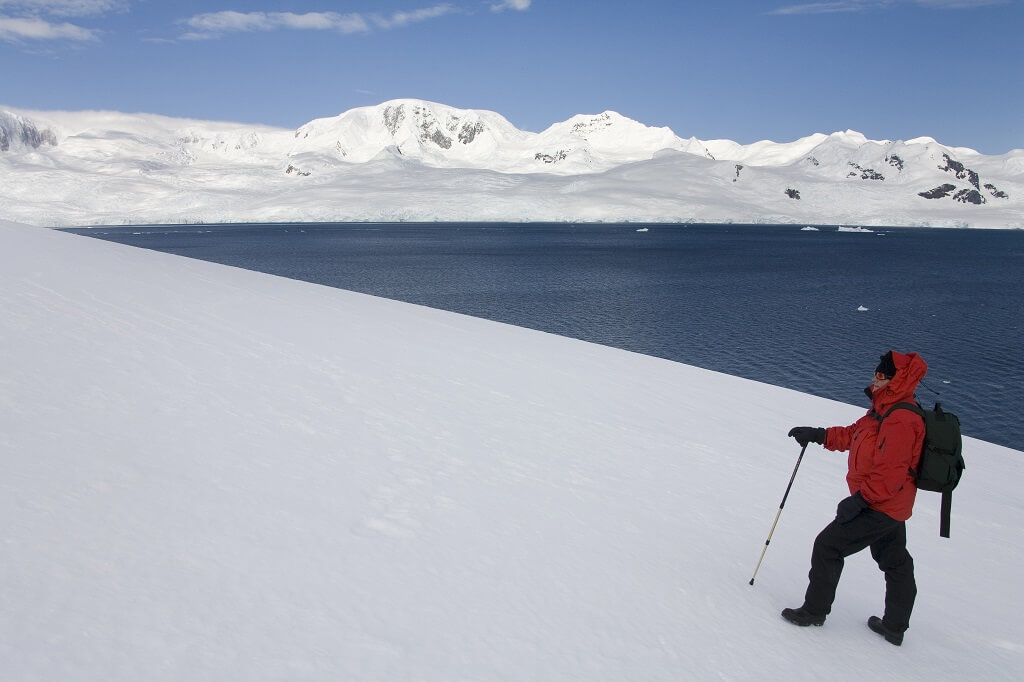 Solo explorer hiking across Antarctic snowfield with coastal mountains and icy ocean in background