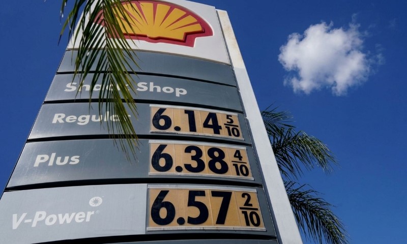 Gas station sign under blue sky showing high prices: Regular $6.14, Plus $6.38, V-Power $6.57. Palm frond and fluffy cloud in background