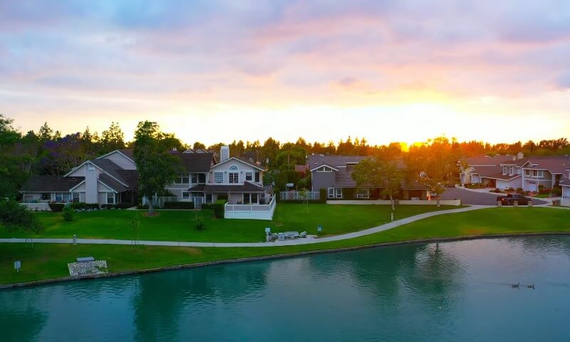 Suburban neighborhood at sunset with houses along a tranquil lake, lush green lawns, and a vibrant sky