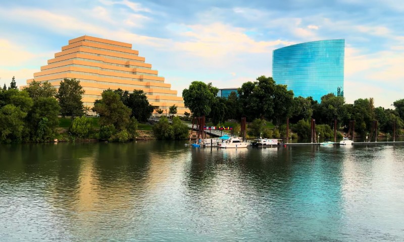 Pyramid-shaped beige building and a curved blue glass tower by a calm river