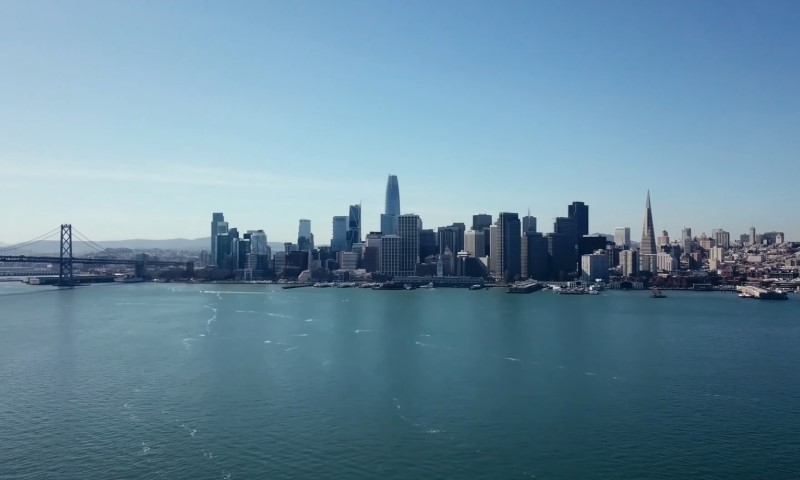 A calm San Francisco skyline with modern skyscrapers under a clear blue sky is reflected in a serene bay