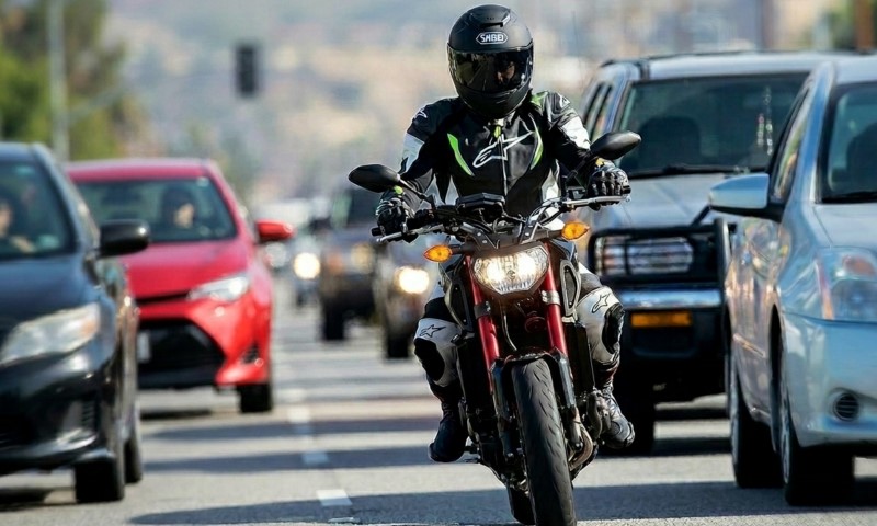 A motorcyclist in black gear and helmet rides on a busy city street, flanked by cars