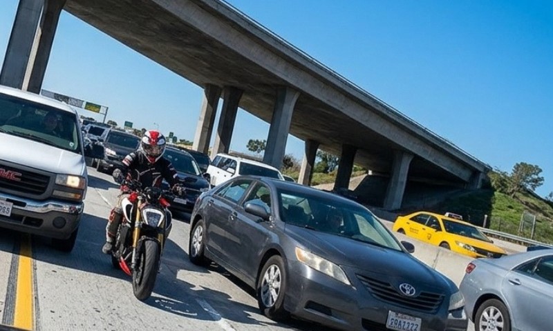 A motorcyclist wearing red and black gear rides between lanes of slow-moving cars on a busy highway