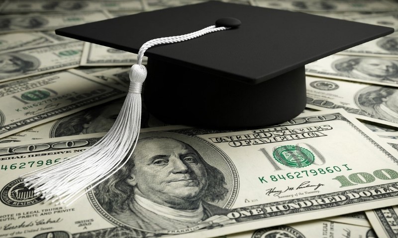 A black graduation cap with a white tassel lies on a bed of one hundred dollar bills