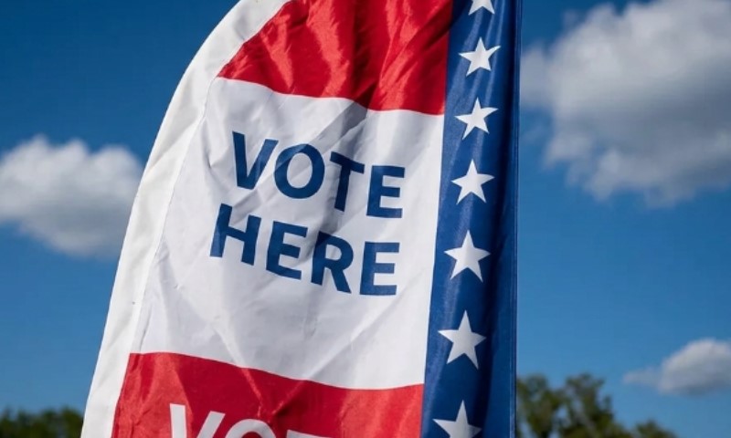 A flag with the words "VOTE HERE" printed on it is displayed against a blue sky. The flag has red and white stripes and a blue section with white stars