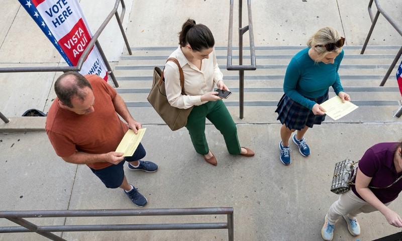 A group of people are standing in line on stairs, holding papers, with a "Vote Here" banner visible