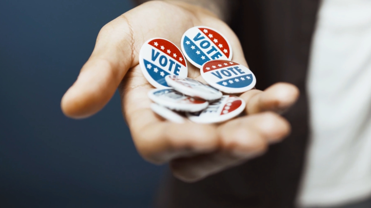 A hand holds several red, white, and blue Vote buttons
