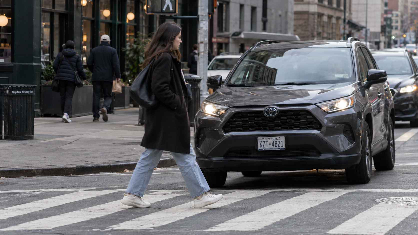 Woman crosses a city crosswalk in front of a gray Toyota SUV with its headlights on.