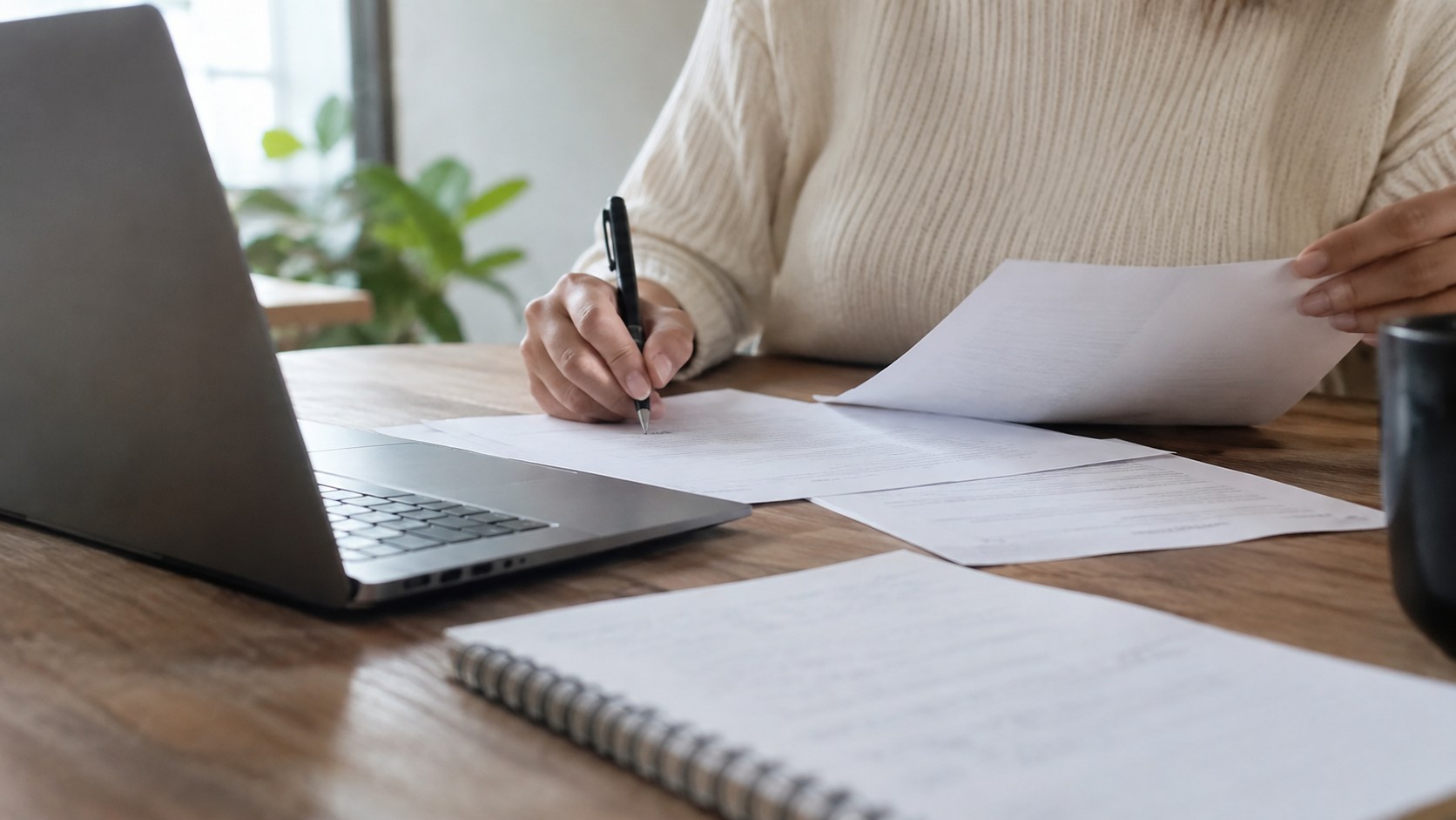 Person reviews and signs business documents beside a laptop.
