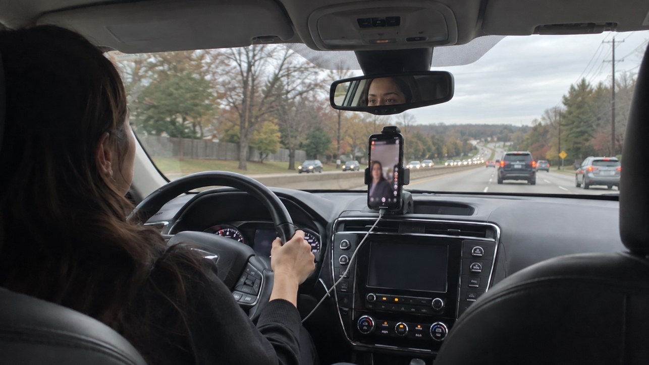A woman drives with a smartphone live stream open on the dashboard.