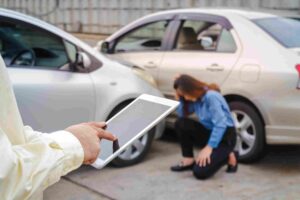 Insurance Companies - Insurance adjuster holding a tablet near two cars involved in a collision while a woman kneels beside the vehicles.