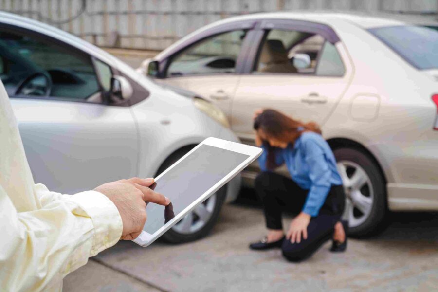 Insurance Companies - Insurance adjuster holding a tablet near two cars involved in a collision while a woman kneels beside the vehicles.