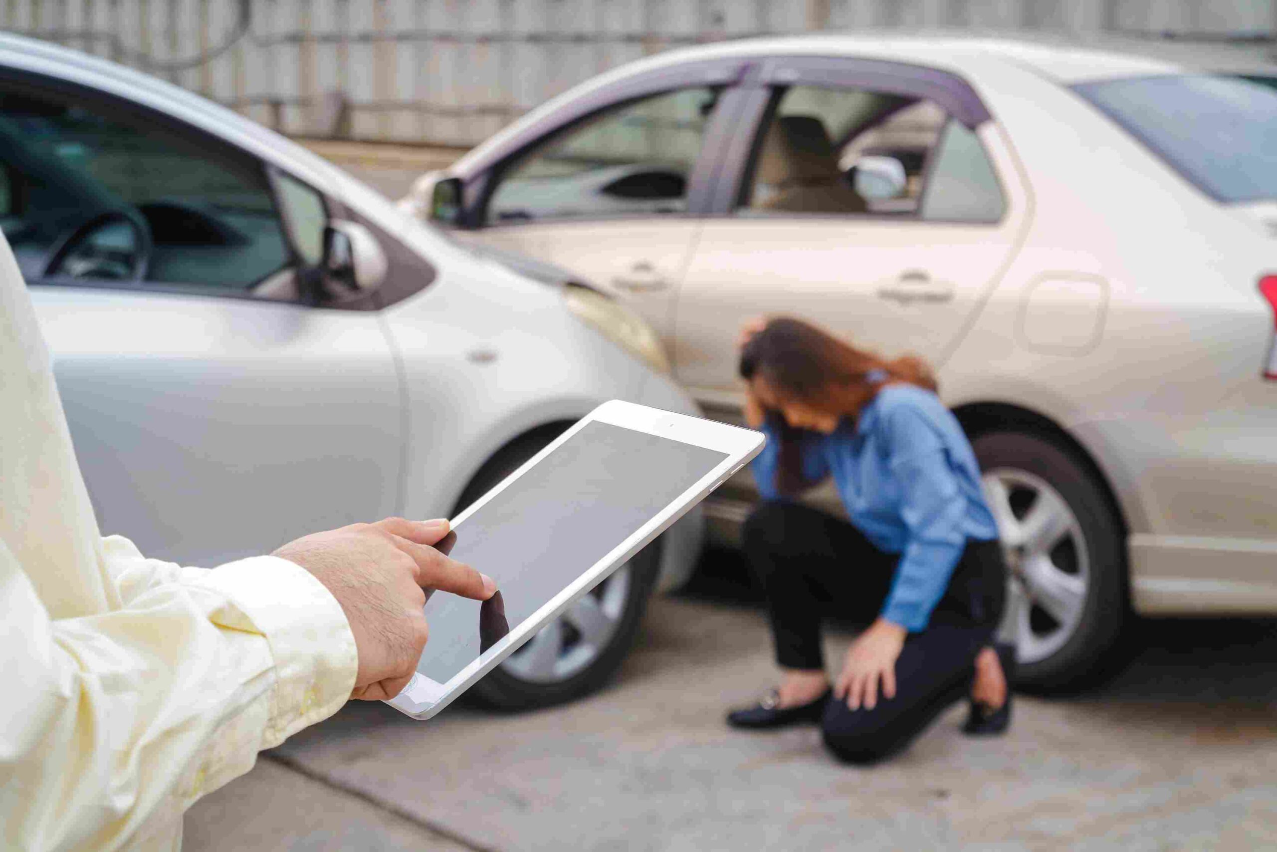 Insurance Companies - Insurance adjuster holding a tablet near two cars involved in a collision while a woman kneels beside the vehicles.