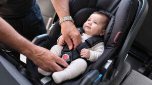 Parent fastening a real baby into a rear-facing child car seat inside a vehicle.