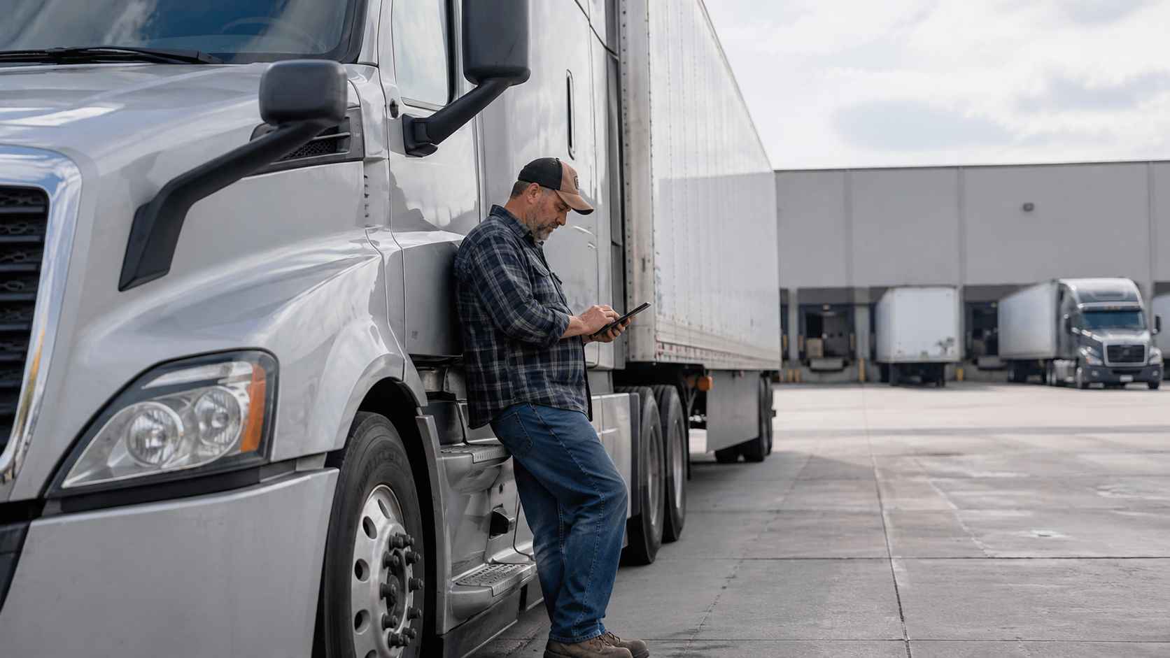 Federal Pilot Program - Truck driver checking a tablet beside a parked semi-truck at a warehouse.