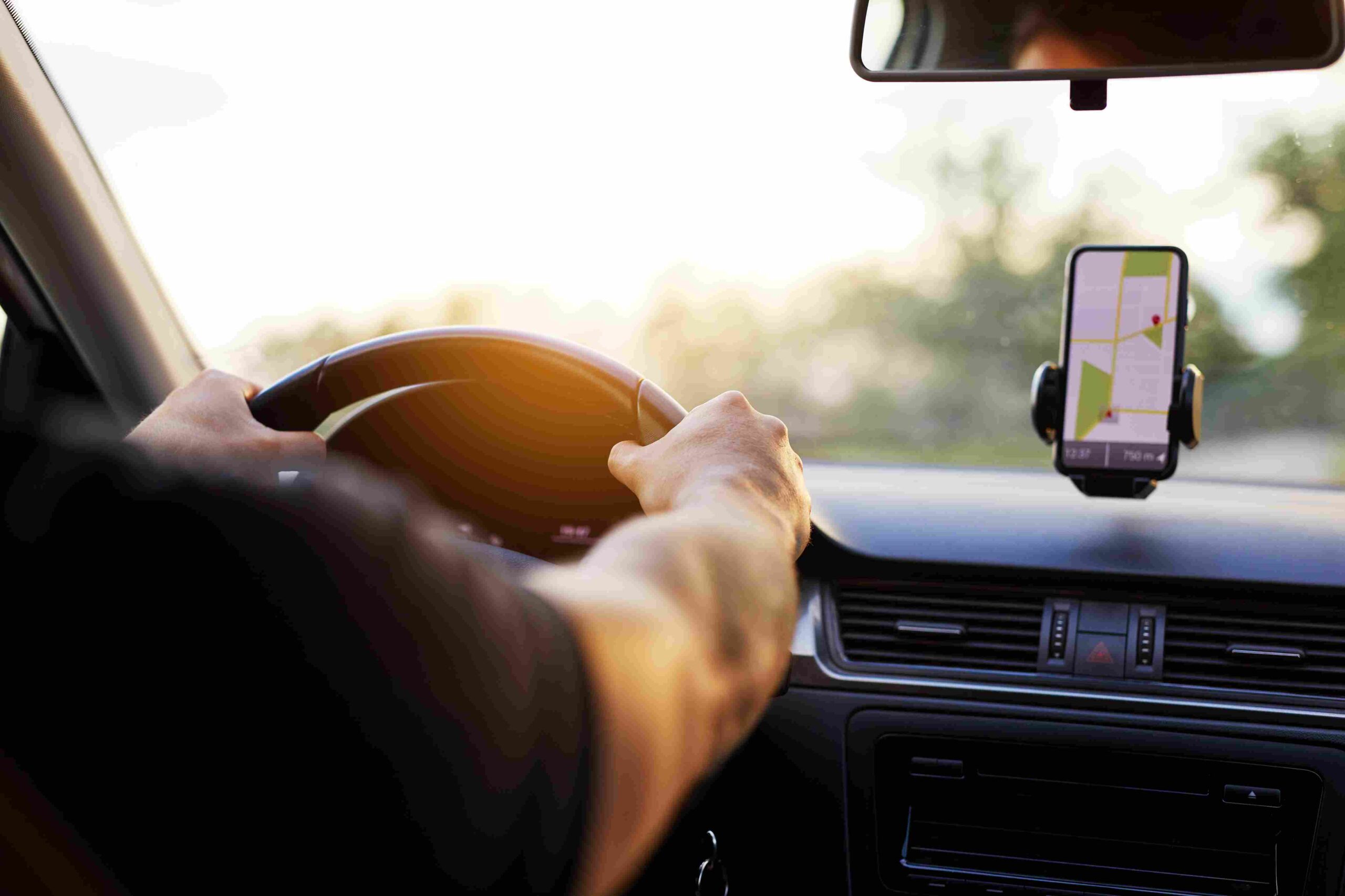 Driver holding a steering wheel with a smartphone displaying GPS navigation on the dashboard.