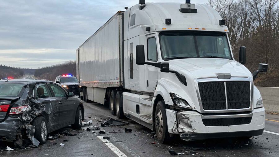 White autonomous-style semi-truck and damaged black sedan at a highway accident scene with police vehicles nearby.