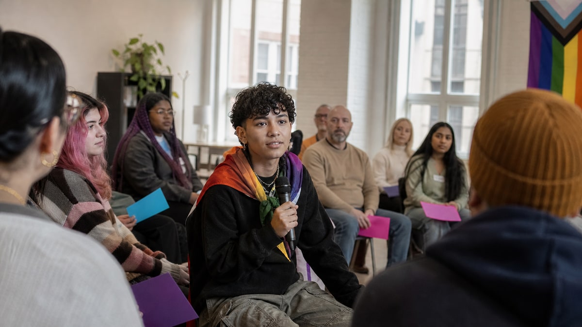 Teen holding a microphone in a colorful community forum with other attendees seated nearby