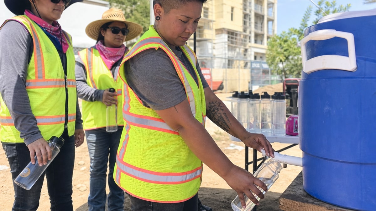 Worker fills a bottle from a blue water cooler at a sunny outdoor worksite