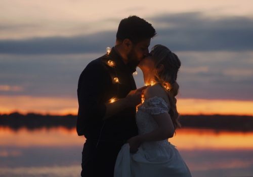 A bride and groom share a kiss at sunset, surrounded by twinkling lights creating a romantic atmosphere