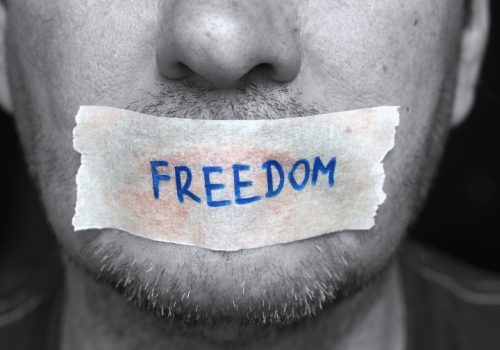 A close-up of a man with tape over his mouth labeled “FREEDOM,” symbolizing restrictions on free speech