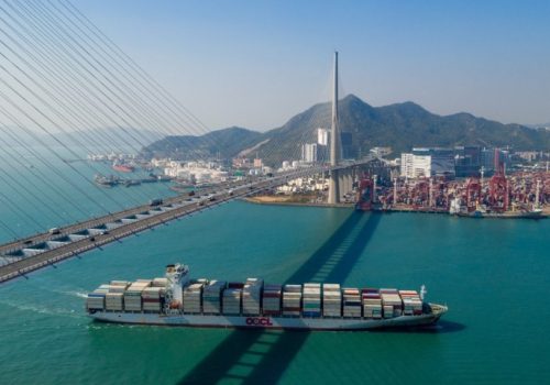 A large container ship sails beneath a suspension bridge in a harbor, with mountains and a busy port in the background