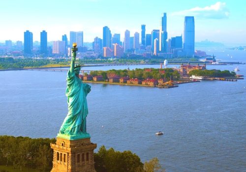 The Statue of Liberty overlooks a calm harbor with boats, lush greenery, and distant skyscrapers under a clear blue sky