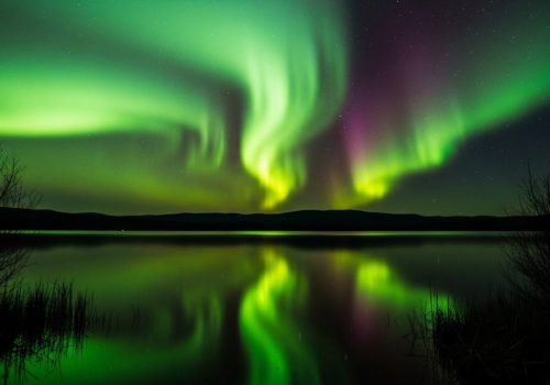 Vivid green and purple northern lights illuminate the night sky, reflecting on a tranquil lake, framed by silhouettes of distant hills and sparse trees