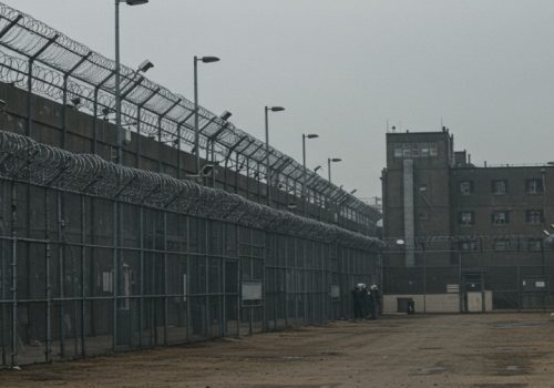 A vast, gray expanse of a correctional facility, enclosed by razor wire and concrete walls