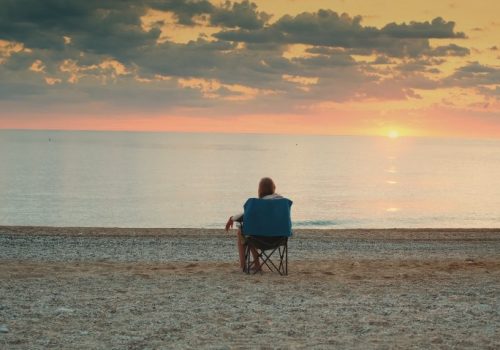 Golden hour paints the sky, while a solitary figure sits on the beach